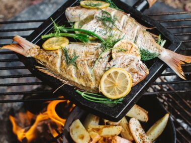 Whole fish and potato wedges cooking in pans over a campfire.