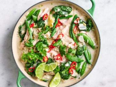 An aerial view of a large green pan filled with green beans, snow peas, and chicken in coconut sauce. Garnished with lime wedges, coriander and red chilli.