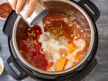 Electric pressure cooker viewed from above with ingredients inside, a can of coconut milk is being poured in