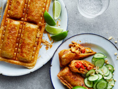 Satay chicken sausage rolls on a plate with cucumber slices, crushed peanuts and lime wedges